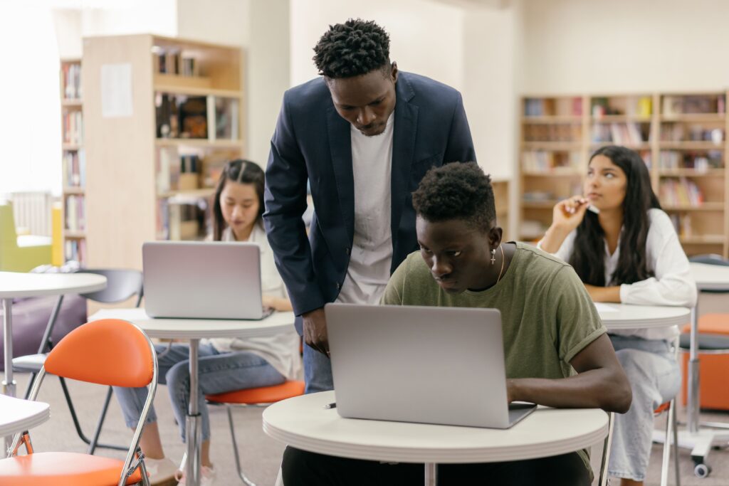 Mentorship Students learning in a classroom setting with a teacher assisting and laptops on desks, creating an interactive education environment.